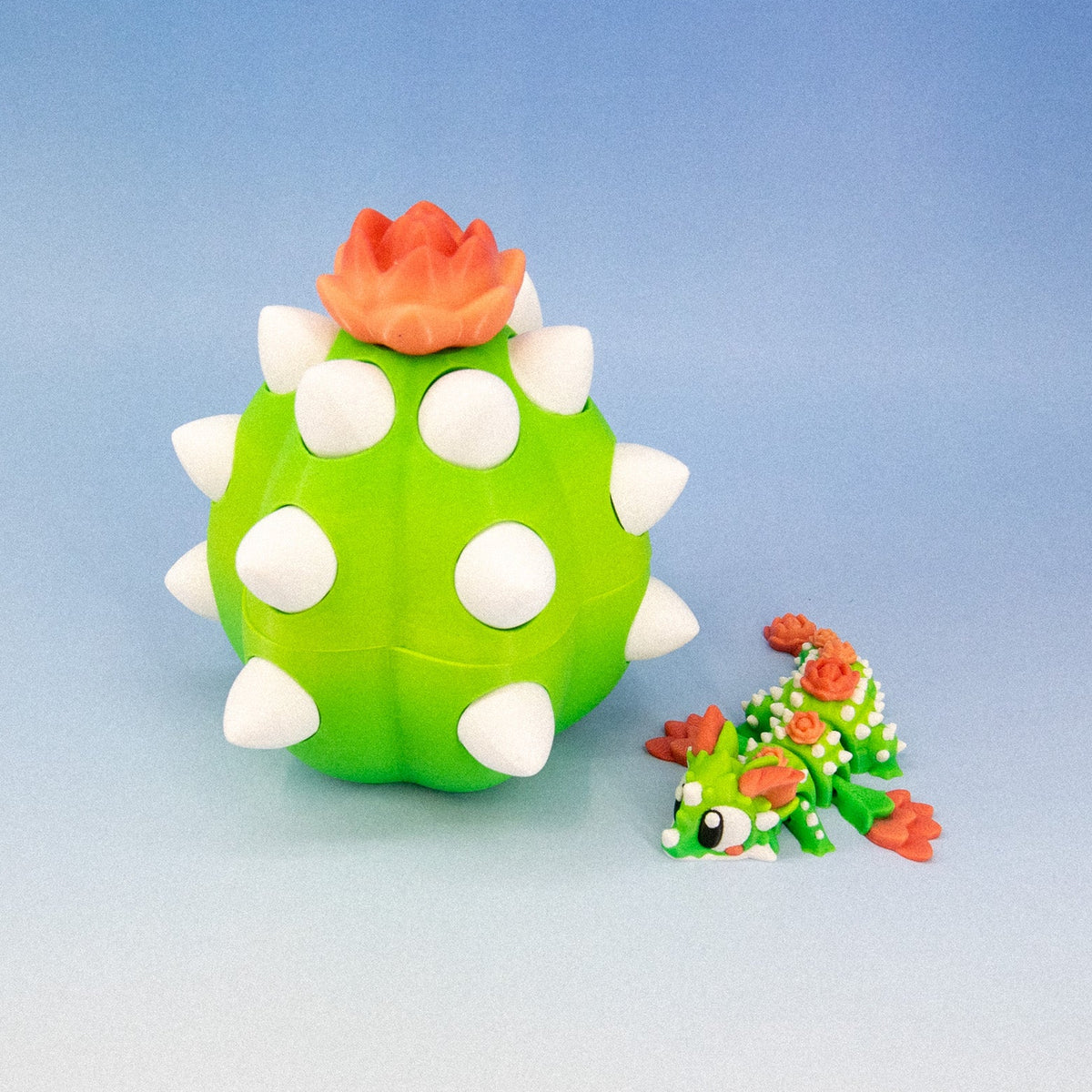 A small green cactus toy with white spikes and an orange flower on top, shown next to a tiny matching creature on a blue gradient background.