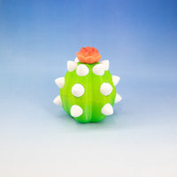 A small cactus-shaped container or toy in bright green with white spikes and a peach-orange flower on top, photographed against a soft blue-to-white gradient background.