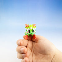 A small green and orange dragon figurine with large white eyes, held in the palm of a hand against a blue gradient background.