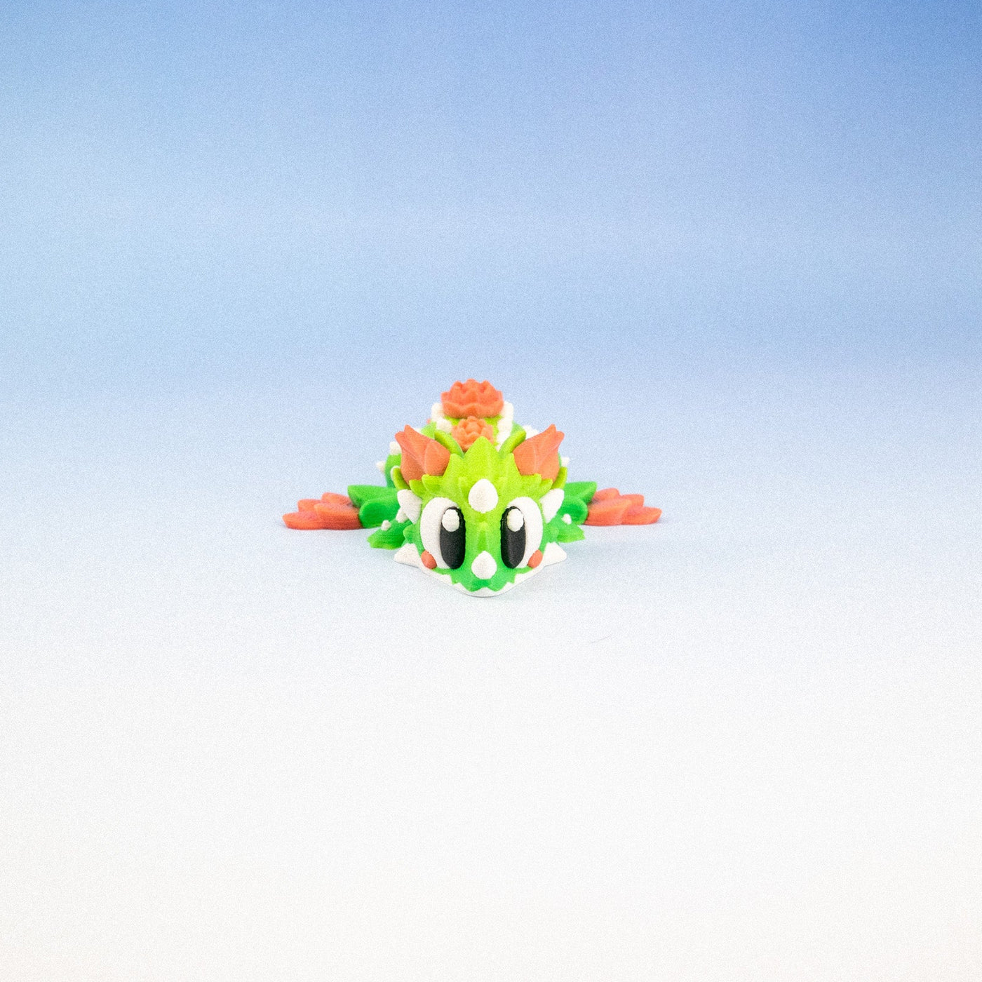 A small green and orange toy dragon figurine with large white eyes, positioned in the lower center of a gradient studio background that transitions from light blue to white.
