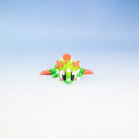 A small green and orange toy dragon figurine with large white eyes, positioned in the lower center of a gradient studio background that transitions from light blue to white.