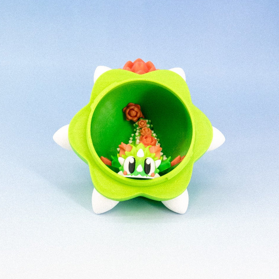A small green star-shaped container or holder with white points and a tiny colorful miniature scene inside, photographed against a blue-to-white gradient studio background.